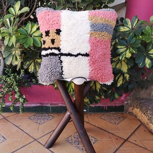 May include: A colorful, geometric patterned throw pillow with pink, white, gray, and yellow accents. The pillow is sitting on a wooden stool with a white seat.