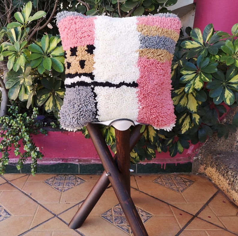 May include: A colorful, geometric patterned throw pillow with pink, white, gray, and yellow accents. The pillow is sitting on a wooden stool with a white seat.