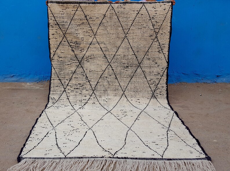 Puede incluir: Una alfombra marroqu&iacute; blanca y negra con un patr&oacute;n de diamantes. La alfombra tiene un borde de flecos.