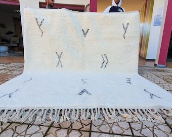 May include: A white Moroccan rug with black geometric patterns. The rug has a fringe border and is laid out on a tiled floor.