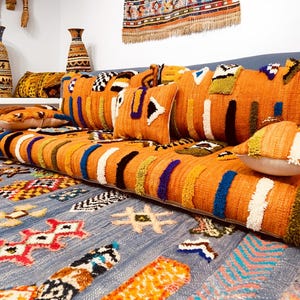 Puede incluir: Un sofá tejido naranja con cojines decorativos y una alfombra a juego. El sofá presenta rayas verticales en varios colores, incluyendo blanco, azul y negro. La alfombra tiene un fondo azul con patrones geométricos coloridos.