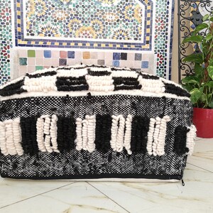 May include: Black and white woven pouf with a geometric pattern. The pouf has a zipper on the side and is made of a soft, fluffy material.