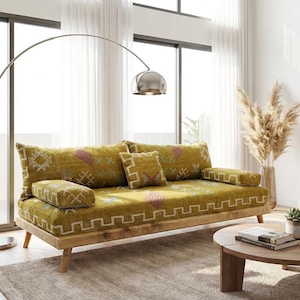 Op de afbeelding: Een woonkamer met een licht houten bank met gele kussens met patronen. Een grote plant in een pot, een ronde salontafel en een decoratief houten wandkunstwerk zijn ook zichtbaar. Een zilveren booglamp zorgt voor verlichting.