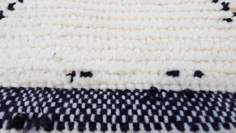 Puede incluir: Una alfombra blanca tejida con un borde a cuadros blanco y negro. La alfombra est&aacute; hecha de un material suave y esponjoso.