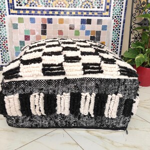May include: Black and white patterned pouf with a woven texture. The pouf has a square shape and is made of thick, fluffy material.