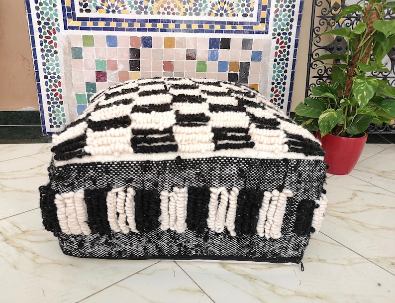 May include: Black and white patterned pouf with a woven texture. The pouf has a square shape and is made of thick, fluffy material.