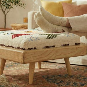 Könnte beinhalten: Ein helles Holz-Ottoman mit einer rechteckigen, gemusterten Oberseite. Die Oberseite hat einen cremefarbenen Stoff mit farbenfrohen geometrischen Designs in Rot, Grün und Blau. Das Ottoman hat vier abgewinkelte Beine und steht auf einem gemusterten Teppich.