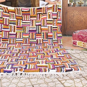 May include: A colorful Moroccan rug with a geometric pattern in shades of orange, purple, white, and black. The rug has a fringe border and is laid out on a tiled floor.