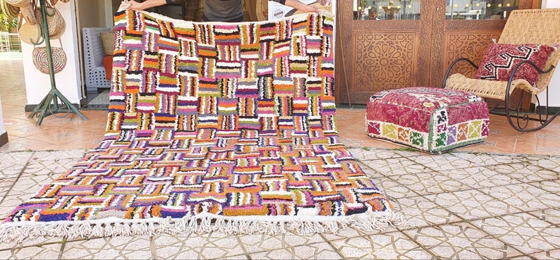 May include: A colorful Moroccan rug with a geometric pattern in shades of orange, purple, white, and black. The rug has a fringe border and is laid out on a tiled floor.