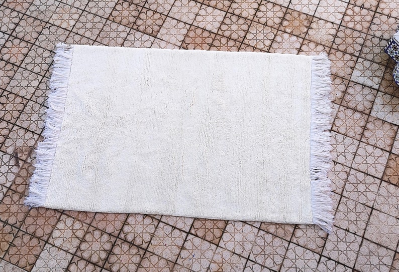 K&ouml;nnte beinhalten: Eine wei&szlig;e Badematte mit Fransenrand. Die Matte liegt auf einem gefliesten Boden.