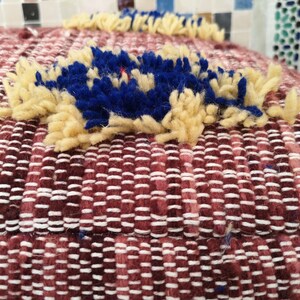 May include: Close-up of a burgundy woven cushion with a blue and cream pom-pom detail.