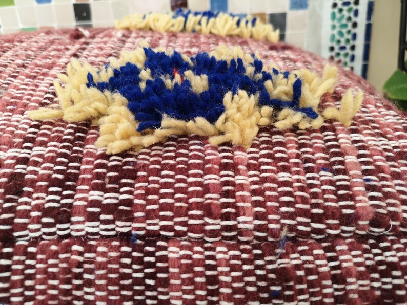 May include: Close-up of a burgundy woven cushion with a blue and cream pom-pom detail.