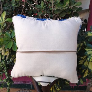 May include: White throw pillow with a zipper closure and blue fringe trim. The pillow is sitting on a wooden chair.