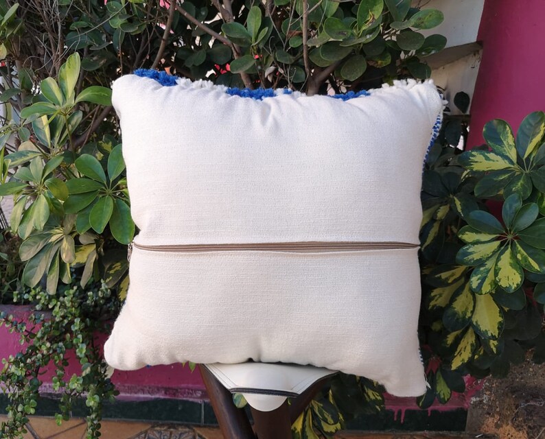 May include: White throw pillow with a zipper closure and blue fringe trim. The pillow is sitting on a wooden chair.