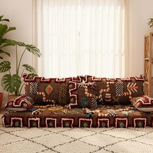 May include: A cozy living room scene featuring a low, brown floor cushion with decorative pillows. The cushion and pillows have colorful geometric patterns. A wooden bookshelf, plants, and a shaggy rug complete the inviting space.