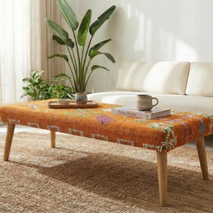 May include: An orange and multi-colored patterned coffee table with wooden legs sits on a woven rug. A white sofa and a large potted plant are in the background. A mug and a book are on the table.