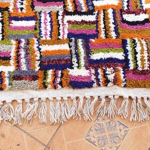 May include: A colorful Moroccan rug with a geometric pattern and fringe. The rug is made of wool and has a variety of colors, including red, orange, yellow, green, blue, and purple.