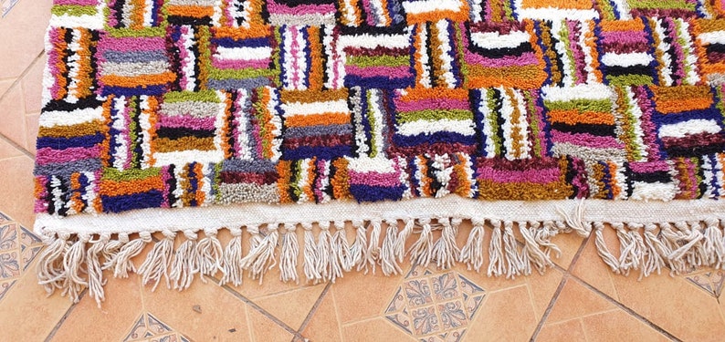 May include: A colorful Moroccan rug with a geometric pattern and fringe. The rug is made of wool and has a variety of colors, including red, orange, yellow, green, blue, and purple.