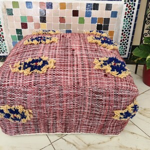 May include: A red and white woven pouf with a cream border and blue and yellow embroidered flowers. The pouf is sitting on a tiled floor.