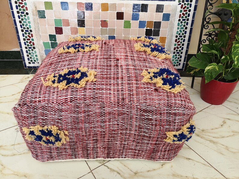 May include: A red and white woven pouf with a cream border and blue and yellow embroidered flowers. The pouf is sitting on a tiled floor.