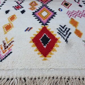 May include: A white rug with colorful geometric patterns in shades of red, yellow, pink, blue, and black. The rug has a fringe border.