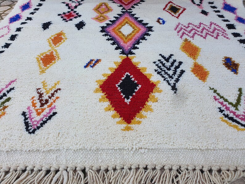 May include: A white rug with colorful geometric patterns in shades of red, yellow, pink, blue, and black. The rug has a fringe border.