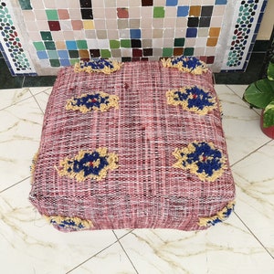 May include: A red and white woven pouf with blue and yellow floral designs. The pouf is a square shape and has a tufted texture.