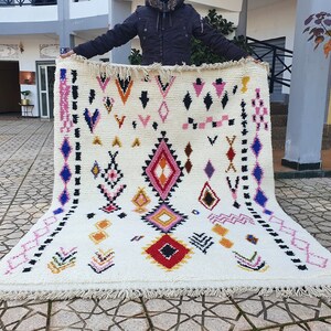 May include: A white wool rug with a geometric pattern in shades of pink, blue, red, yellow, and black. The rug has a fringe border.