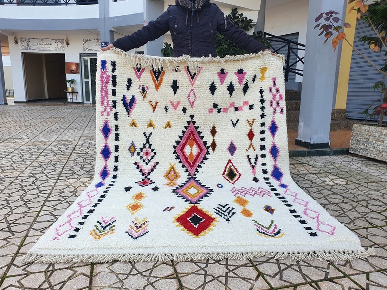 May include: A white wool rug with a geometric pattern in shades of pink, blue, red, yellow, and black. The rug has a fringe border.