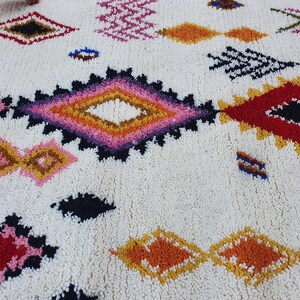 May include: A white rug with a colorful Moroccan-inspired design. The rug features a variety of geometric shapes, including diamonds, squares, and triangles, in shades of red, pink, orange, yellow, and blue.