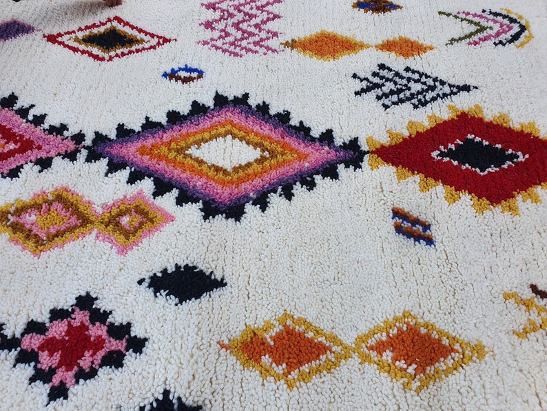 May include: A white rug with a colorful Moroccan-inspired design. The rug features a variety of geometric shapes, including diamonds, squares, and triangles, in shades of red, pink, orange, yellow, and blue.