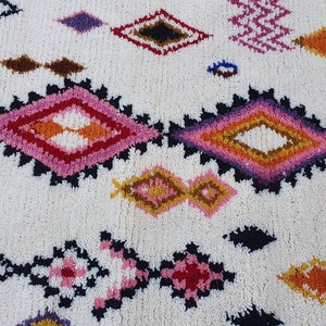 May include: A white rug with a colorful geometric pattern. The pattern features diamonds in shades of pink, orange, yellow, brown, and black.