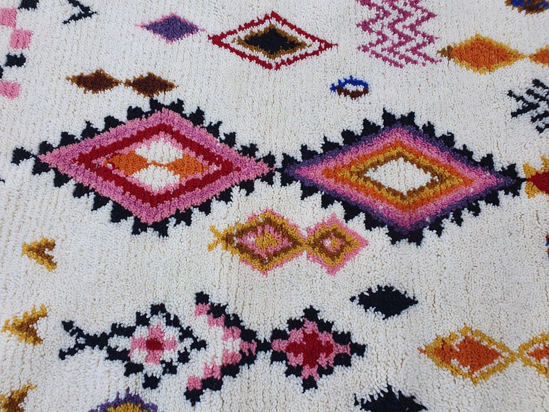 May include: A white rug with a colorful geometric pattern. The pattern features diamonds in shades of pink, orange, yellow, brown, and black.