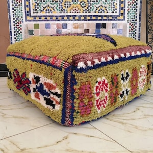 May include: A green, square, tufted ottoman with colorful geometric patterns and fringe details. The ottoman is made of woven fabric and has a soft, plush texture.