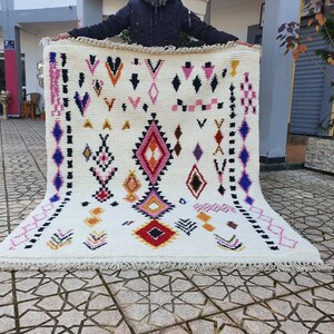 May include: A white wool rug with a geometric pattern in shades of pink, blue, yellow, orange, and red. The rug has a fringe border.