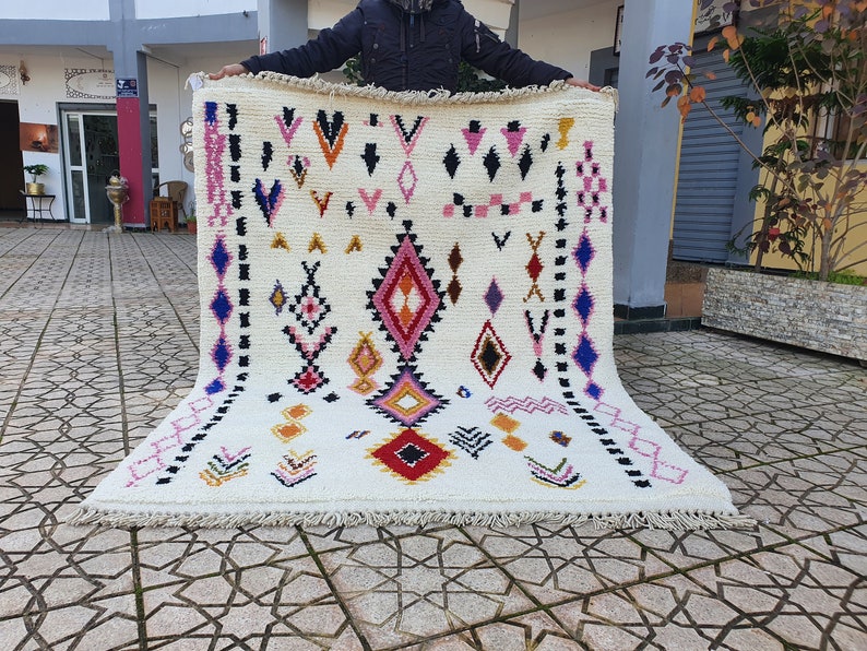 May include: A white wool rug with a geometric pattern in shades of pink, blue, yellow, orange, and red. The rug has a fringe border.