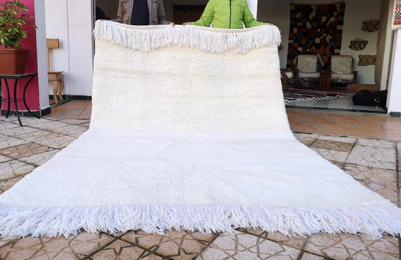 K&ouml;nnte beinhalten: Ein wei&szlig;er, flauschiger Teppich mit Fransenrand. Der Teppich wird von zwei Personen gehalten.