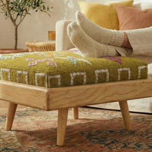 May include: A square ottoman with a light wood frame and four tapered legs. The cushion is a textured green fabric with a colourful floral pattern. The ottoman is placed on a patterned rug.