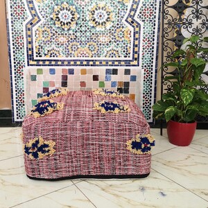 May include: A red and white woven pouf with blue and yellow tassels. The pouf is sitting in front of a tiled wall with a geometric pattern.