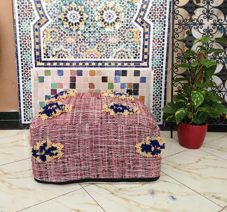 May include: A red and white woven pouf with blue and yellow tassels. The pouf is sitting in front of a tiled wall with a geometric pattern.