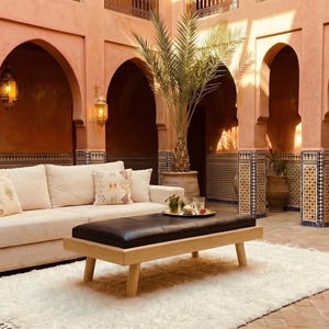 Könnte beinhalten: Ein beiges Sofa mit mehreren Kissen, eine schwarze Leder-Ottomane und ein weißer Teppich befinden sich in einem Innenhof mit gewölbten Türen. Die Wände sind terrakottafarben und der Boden ist gefliest. Dekorative Laternen hängen an den Wänden.