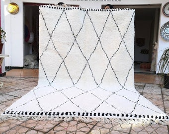 May include: A white Moroccan rug with a black diamond pattern. The rug has a fringe border and is made of wool.
