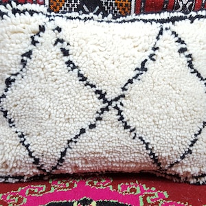 May include: White and black Moroccan pouf pillow with a diamond pattern. The pillow is made of fluffy wool and has a fringe border.