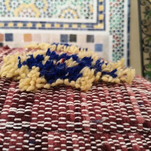May include: A close-up of a red and white woven cushion with a yellow and blue fringe detail. The cushion is made of a soft, textured fabric.