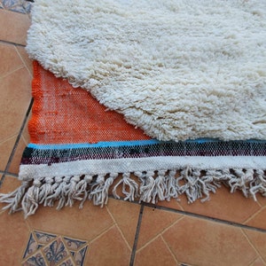 Op de afbeelding: Een wit en oranje kleed met een franje rand. Het kleed heeft een witte, pluche textuur en een geweven oranje en blauw patroon.