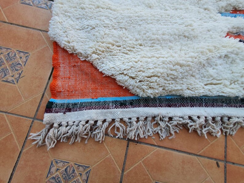 Op de afbeelding: Een wit en oranje kleed met een franje rand. Het kleed heeft een witte, pluche textuur en een geweven oranje en blauw patroon.