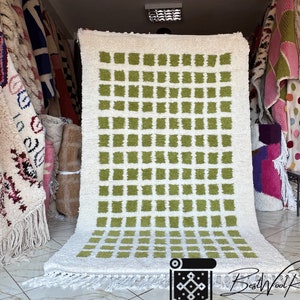 May include: A white and green Moroccan rug with a geometric pattern of squares. The rug has a fringe border and is displayed in a shop.