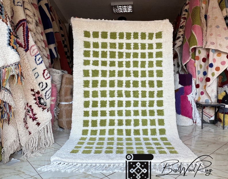 May include: A white and green Moroccan rug with a geometric pattern of squares. The rug has a fringe border and is displayed in a shop.
