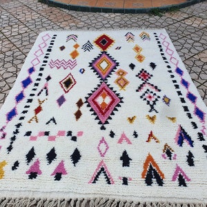 May include: A white Moroccan rug with a geometric pattern in shades of pink, blue, black, orange, and red. The rug has a fringe border.