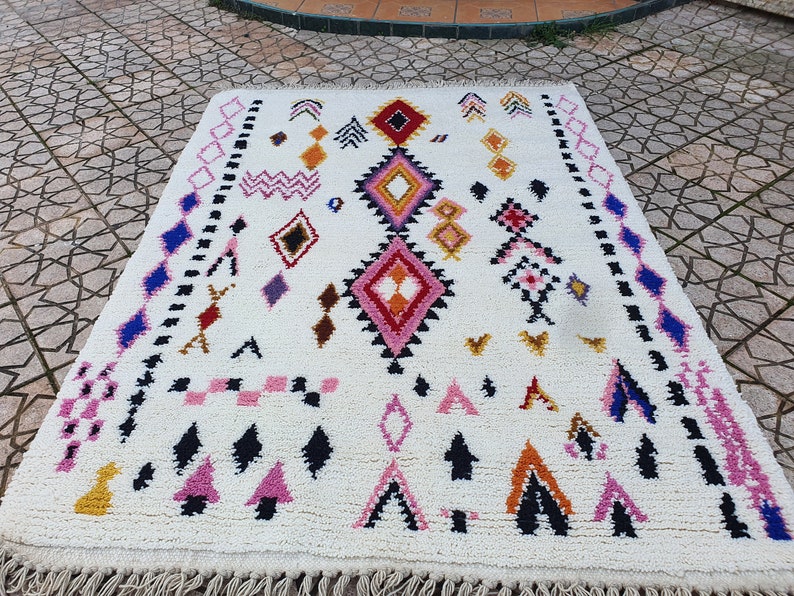 May include: A white Moroccan rug with a geometric pattern in shades of pink, blue, black, orange, and red. The rug has a fringe border.